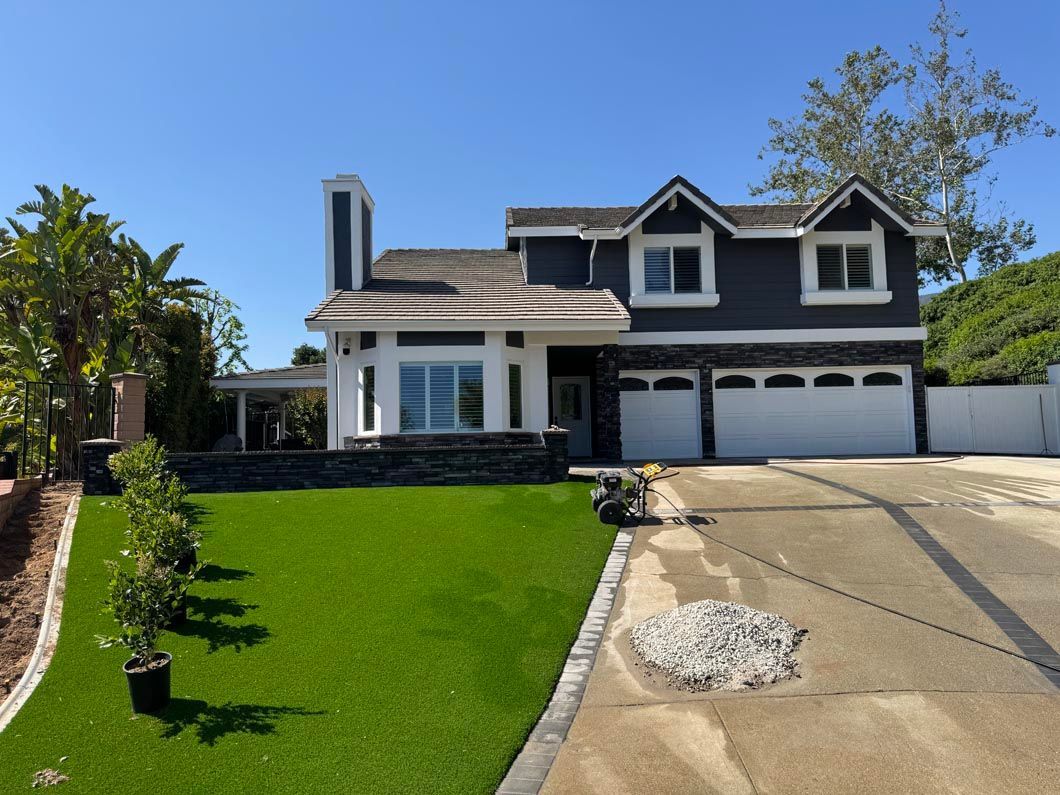 Two-story dark house with white trim, green lawn, and sloped driveway under a clear blue sky