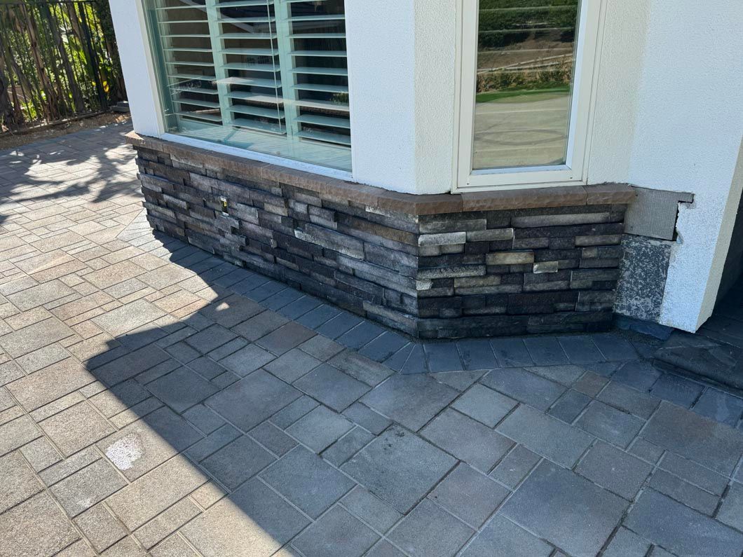 Corner of a building with stacked-stone base, white wall, and gray paver walkway in sunlight