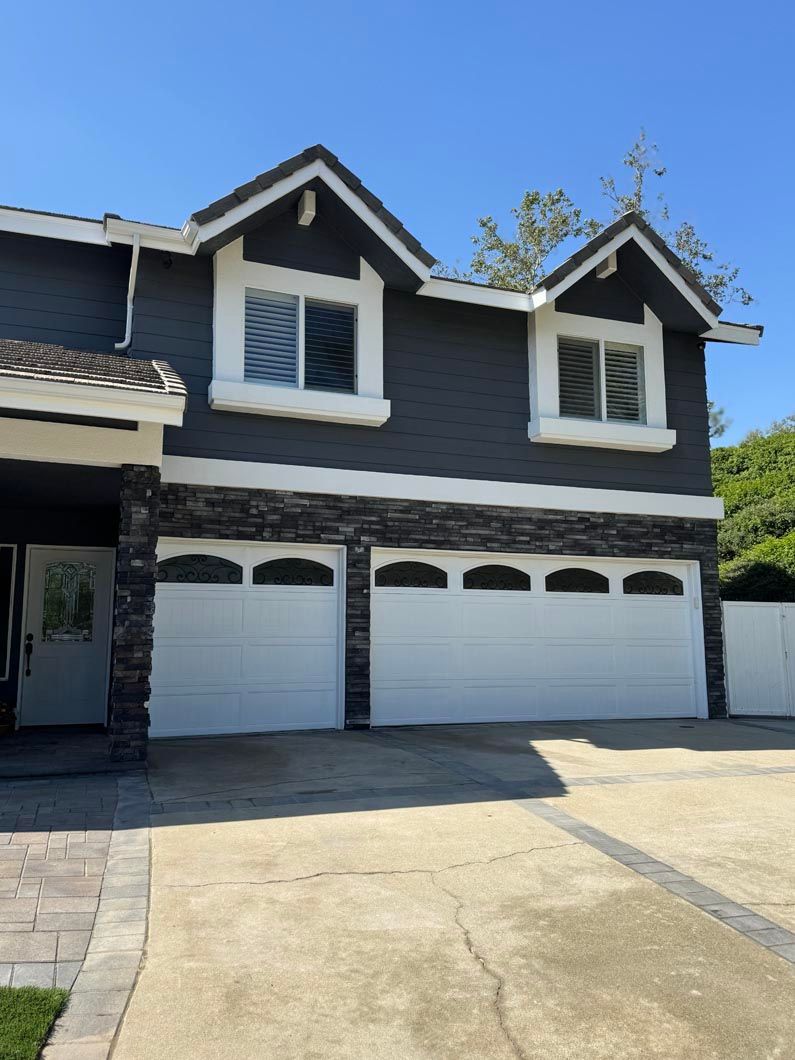 Two-story dark gray house with white trim and three white garage doors under a bright blue sky