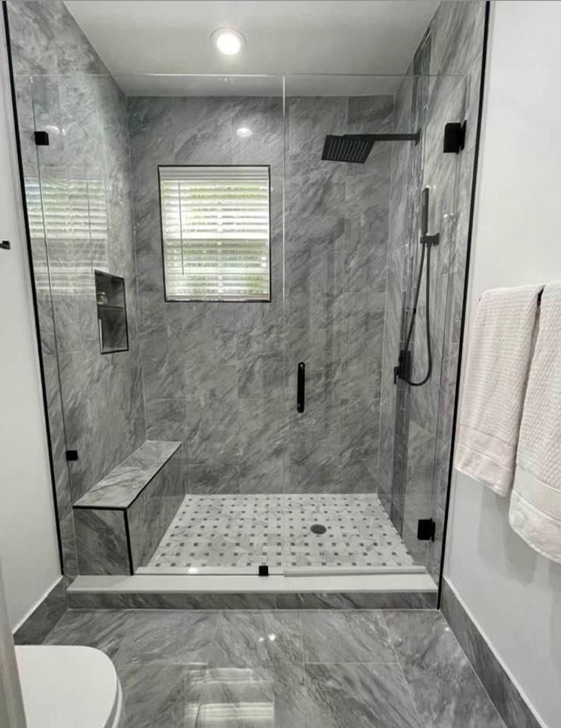 Modern gray-tile walk-in shower with glass door, bench, showerhead, window, and towels on the wall