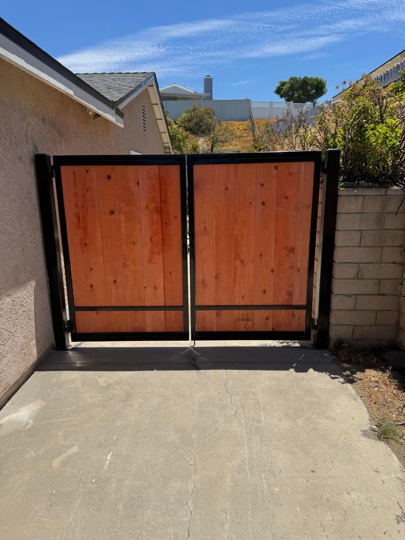 Wooden double gate with black frame at the end of a concrete driveway, beside a brick wall.