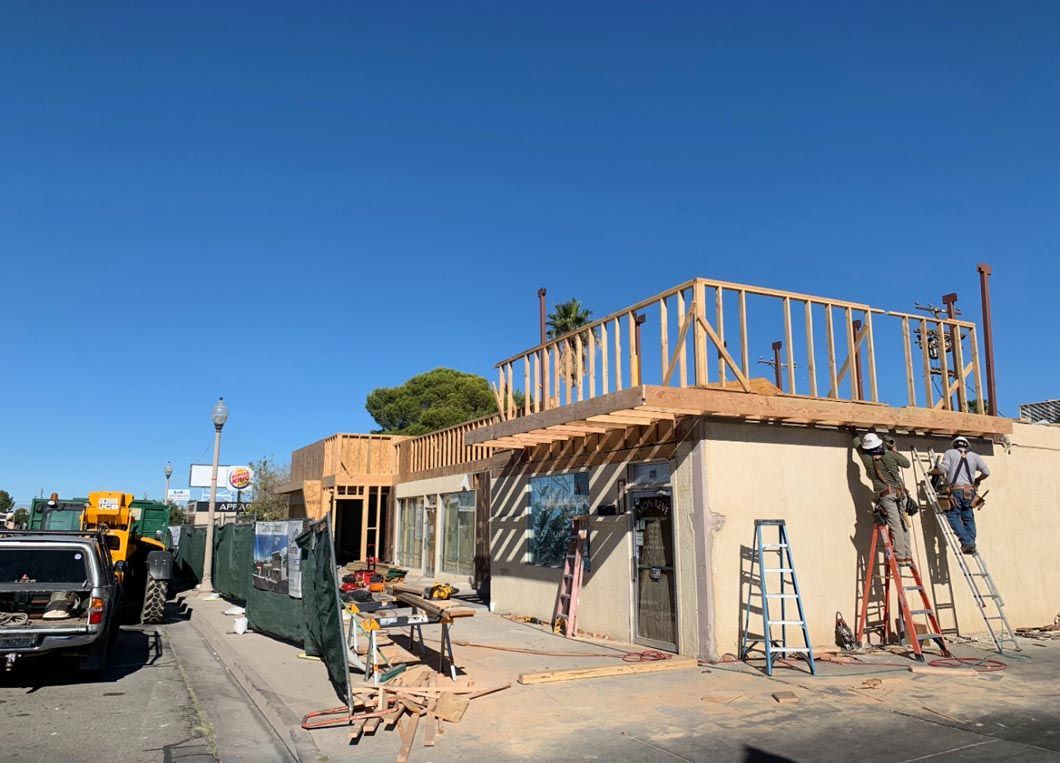 Two-story building under construction on a sunny street, with scaffolding, ladders, and parked vehicles nearby