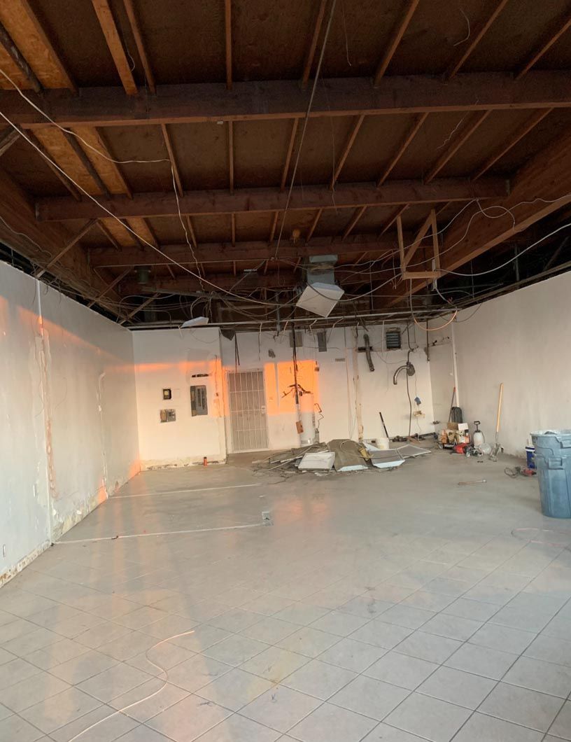 Empty unfinished room with exposed ceiling beams, white walls, and tiled floor lit by orange window light