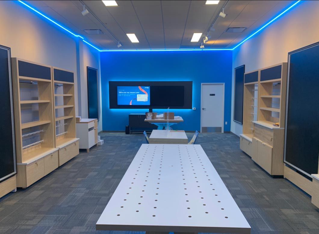 Blue-lit classroom or lab with shelves, tables, and a wall-mounted screen at the far end.