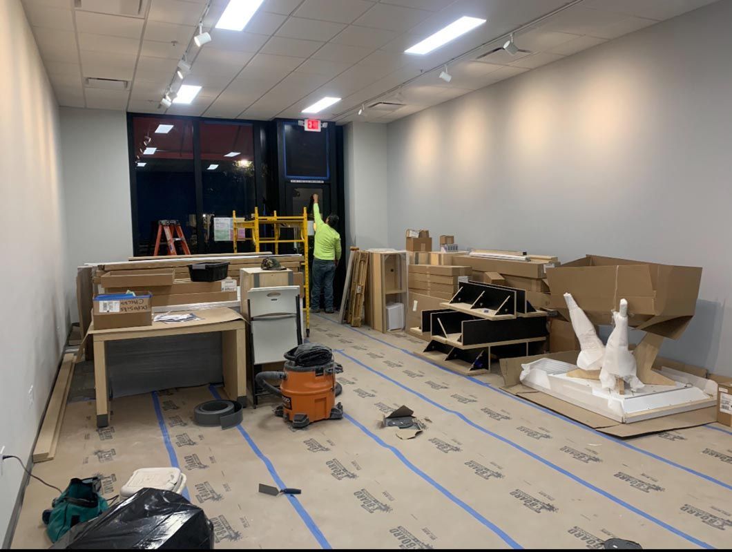 Long unfinished room with cardboard, boxes, tools, and wrapped items under bright ceiling lights
