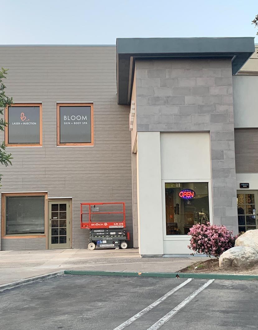 Storefronts in a strip mall with gray siding, orange-trimmed windows, and a pink flowering bush by the entrance