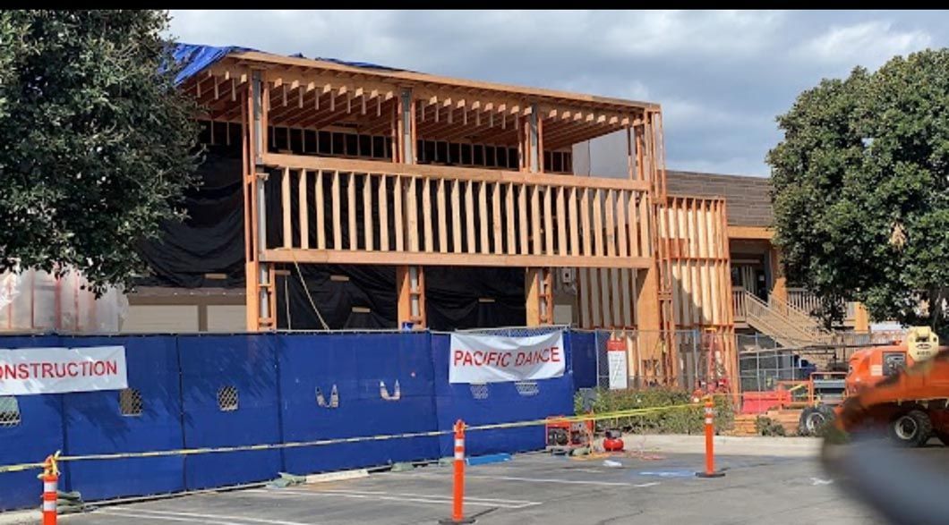 Wooden building under construction behind a blue fence, with orange cones and equipment on the street.