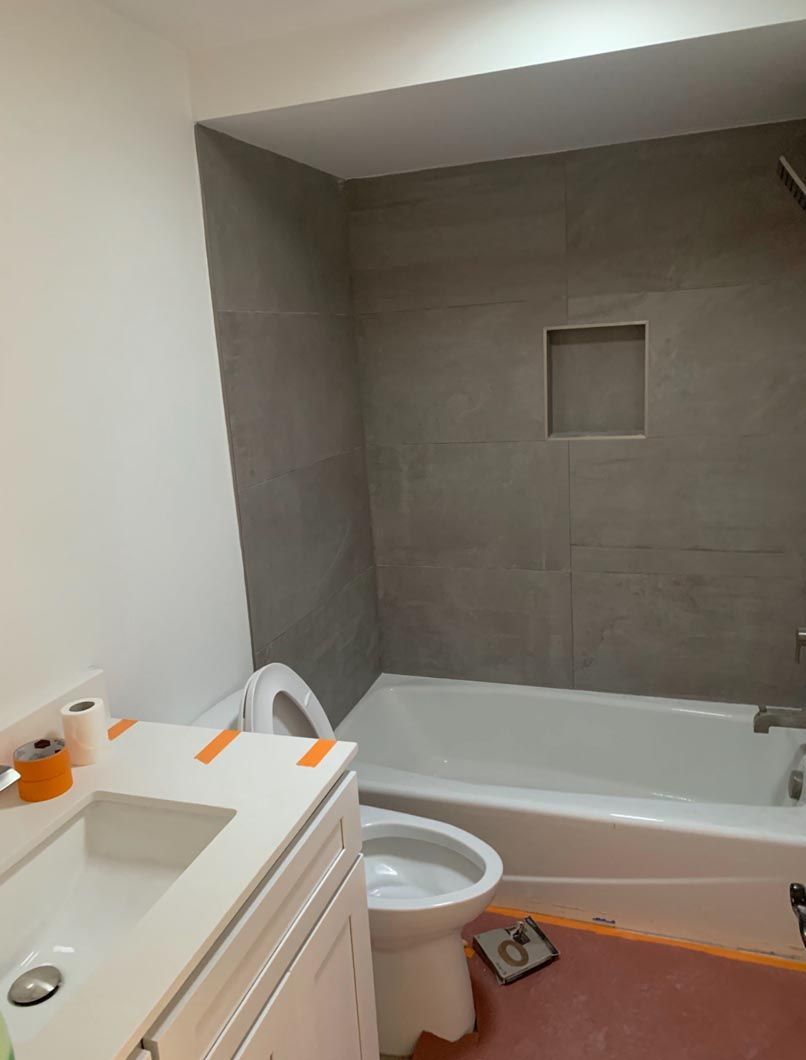 Small bathroom with a white sink and toilet beside a tub, gray tiled shower wall, and orange floor covers.