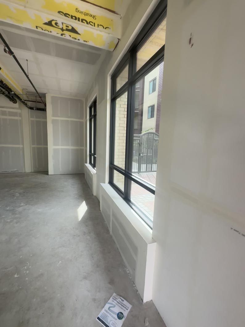 Empty unfinished room with gray floor, white walls, large windows, and exposed ceiling beams.