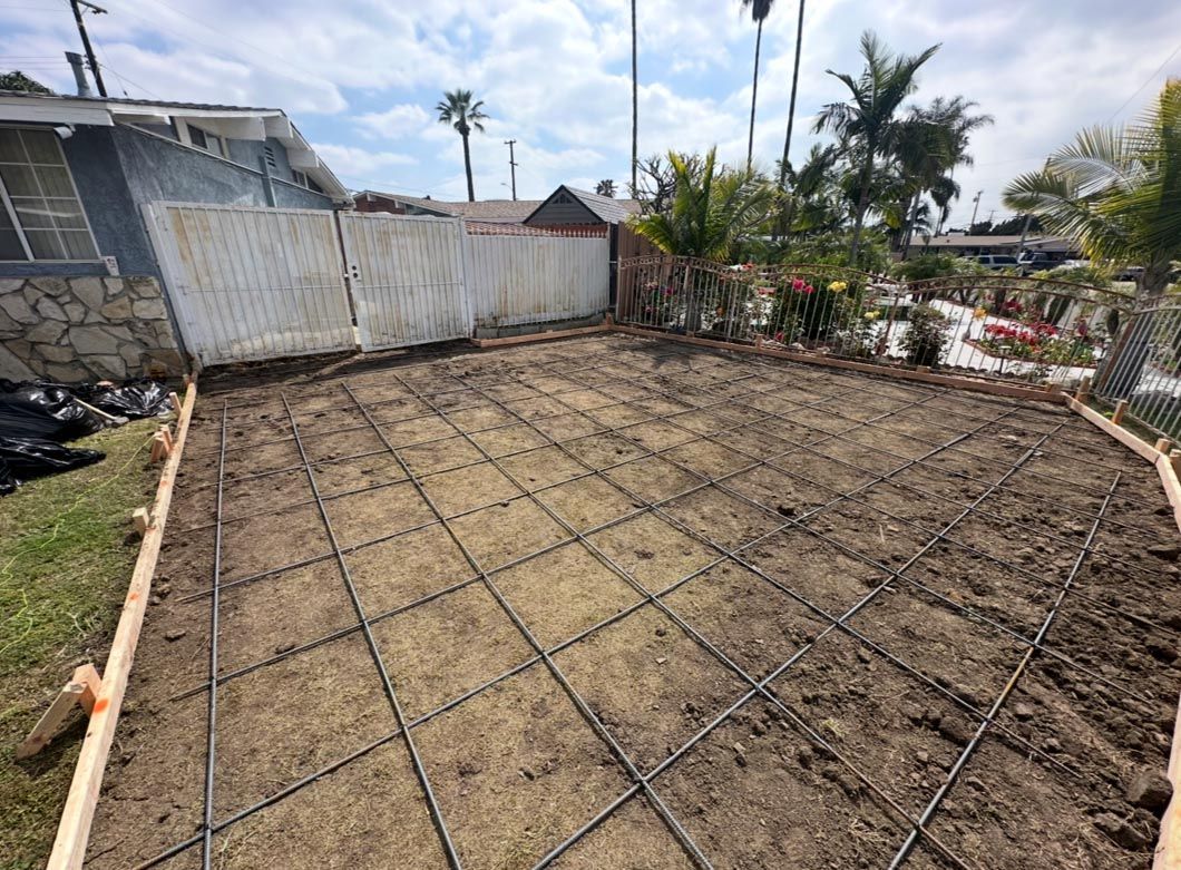 Construction site with rebar mesh laid over dirt beside a white fence and palm trees