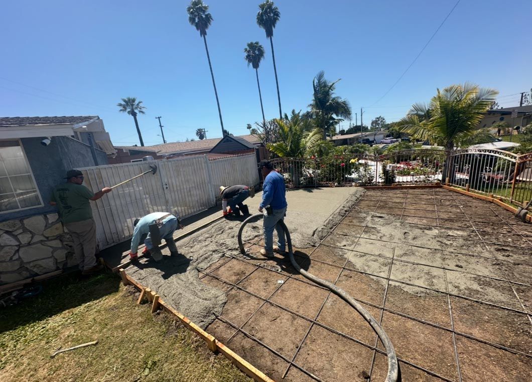 Two workers smoothing wet concrete in a backyard, with a wheelbarrow and fence nearby.