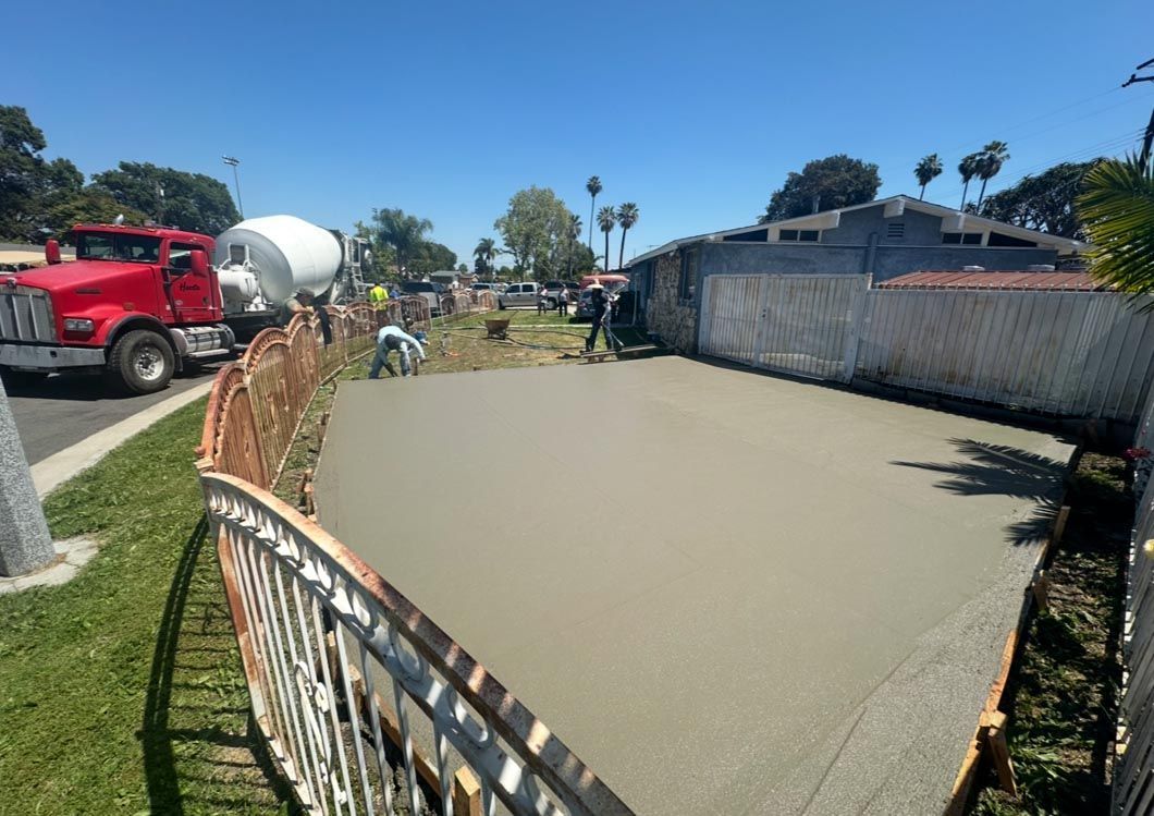Concrete pool under construction beside a red cement truck, with workers and fencing on a sunny day