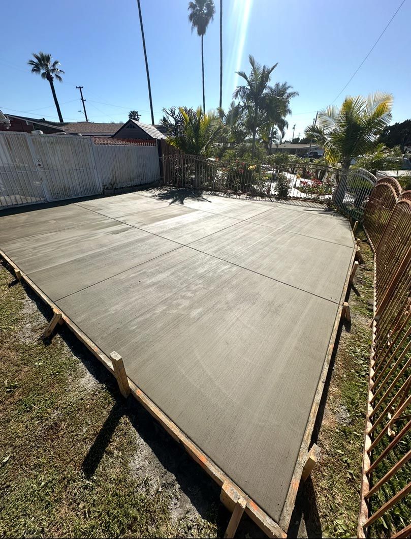 Freshly poured concrete patio slab beside a chain-link fence and palm trees in a sunny yard