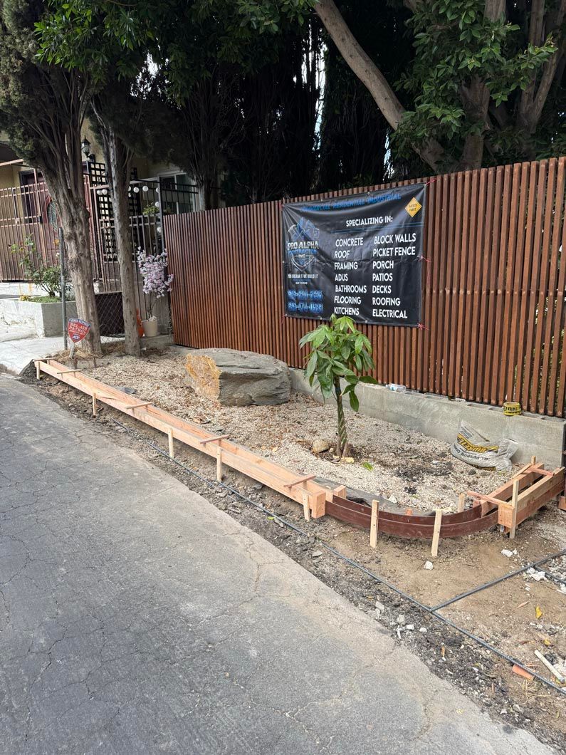 Small fenced roadside garden with a young tree and a sign beside a sidewalk and wall.