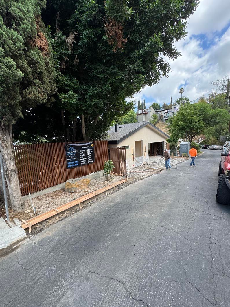 Street with a small building, trees, construction debris, and a person in an orange shirt walking away.