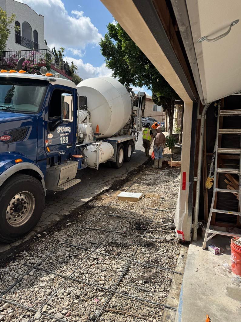 Blue cement mixer truck on a narrow gravel street beside a building and sidewalk