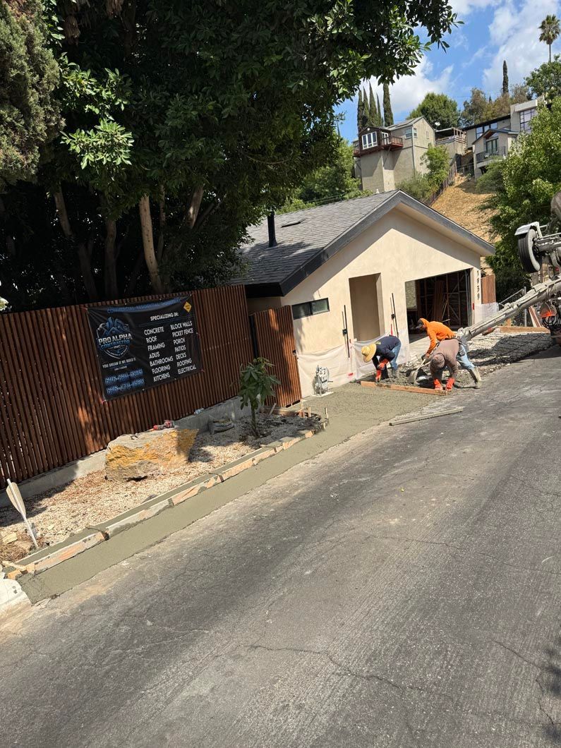 Roadside construction scene with a small beige shed, workers, a fence, and trees on a sloped street.