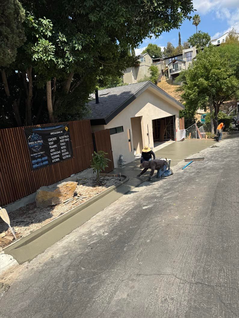 Person sitting on a bench outside a small beige building on a sunny hillside street.