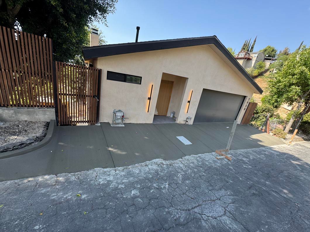 Cream-colored house with garage, sloped driveway, and wooden gate beside gravel and trees