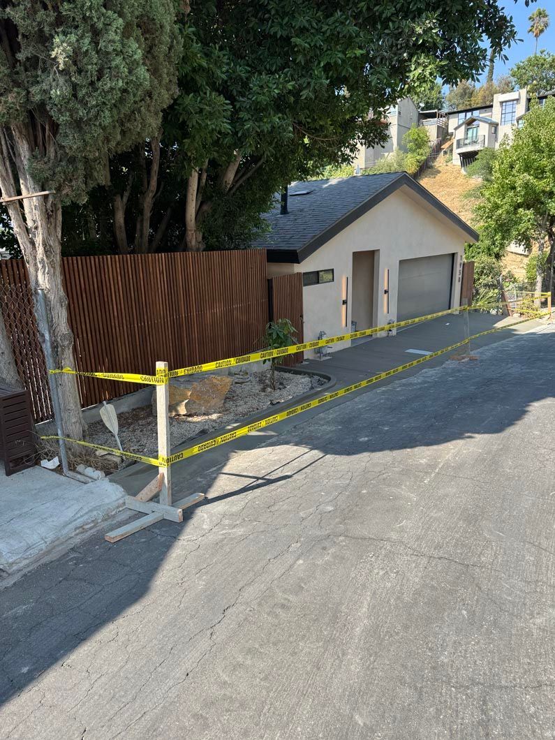 Residential driveway with yellow caution tape, beige garage, and large trees on a sloped street