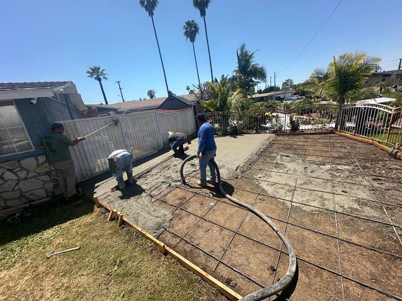 Worker pressure-washing a concrete yard beside a stucco wall and garden under a sunny sky
