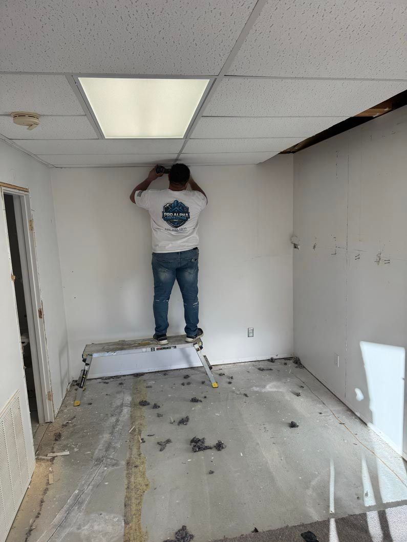 Person standing on a ladder in a bare room, working on the ceiling near a fluorescent light.