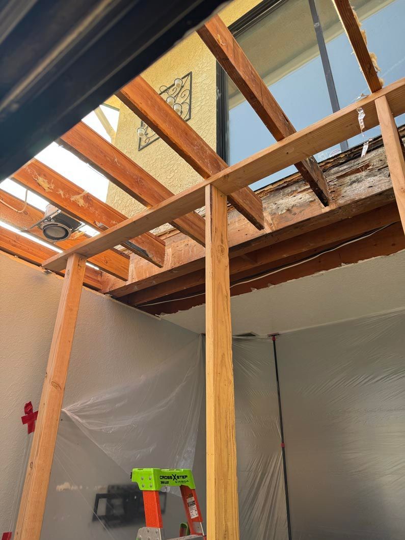 Construction area with exposed wooden ceiling joists, plastic sheeting, and a ladder below.
