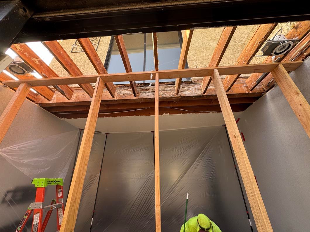 Exposed wooden ceiling framing in a room under renovation, with a ladder on the left and worker below.