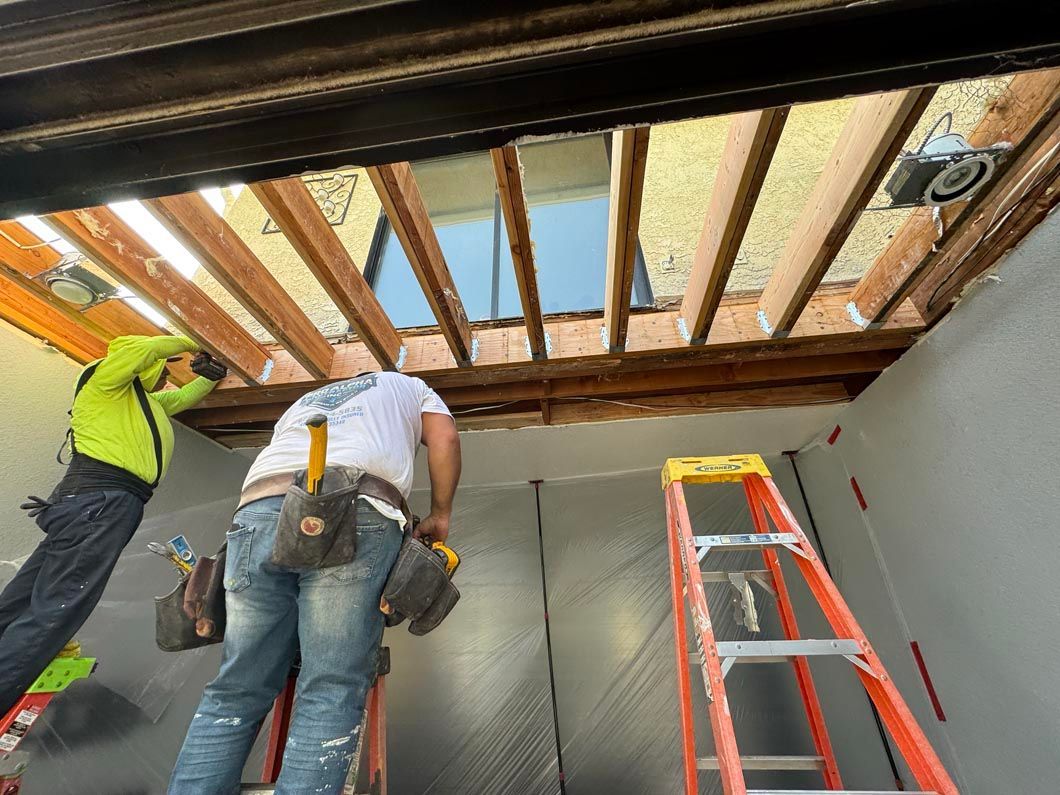 Two workers installing ceiling insulation in a room, with a ladder and exposed beams visible.