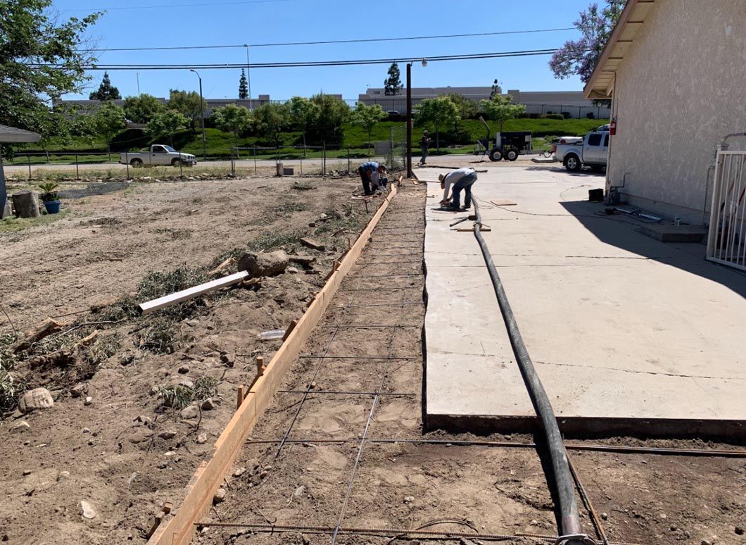 Backyard concrete slab beside a dirt strip and exposed pipes under a bright blue sky