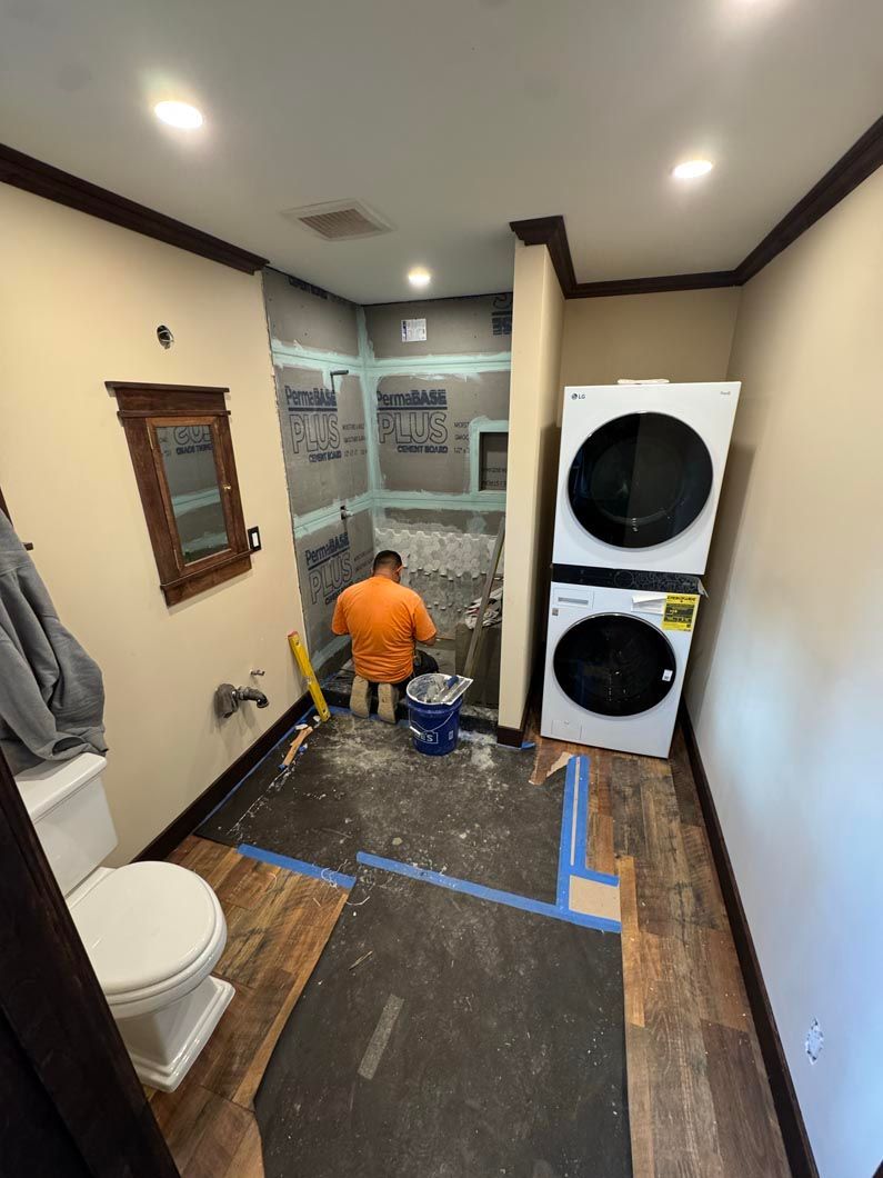 Worker kneeling in a narrow bathroom during renovation, with floor covered and stacked washer and dryer nearby