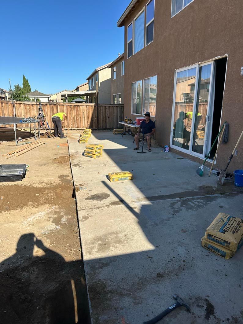 Backyard patio construction with workers, concrete slabs, tools, and a two-story house under a blue sky