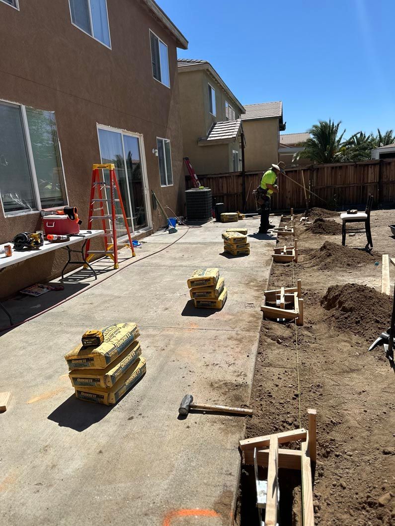 Backyard construction site with concrete slabs, tools, ladder, and a worker near a fence.