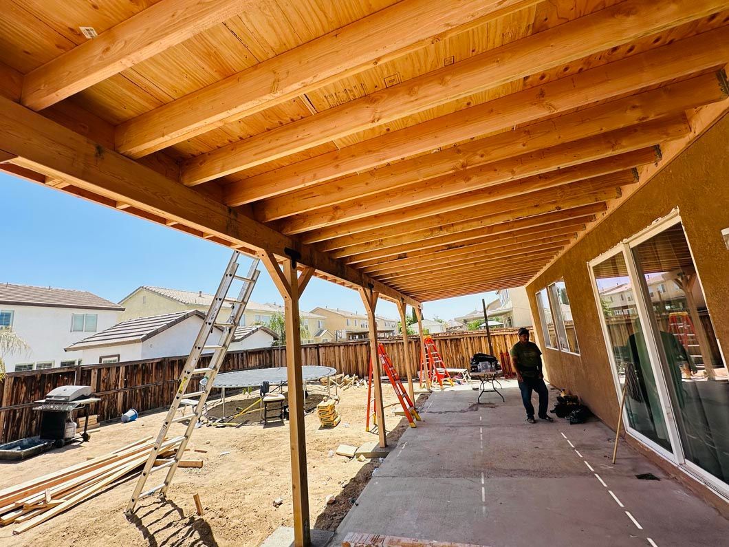 Covered patio under construction with exposed wood beams, ladder, and workers beside a house