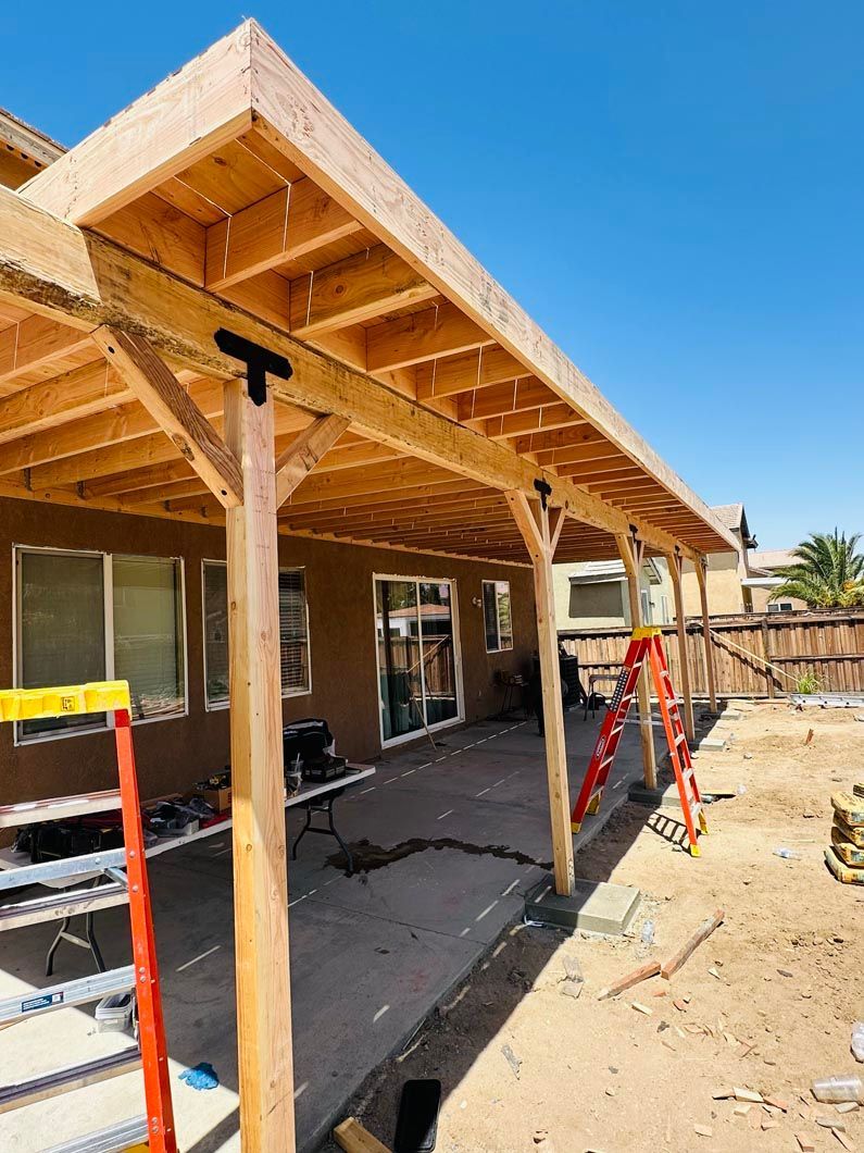 Partially built wooden patio roof with red supports beside a house under a blue sky