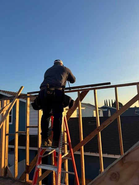 Person on a ladder working on a wooden rooftop frame at a construction site under a clear blue sky