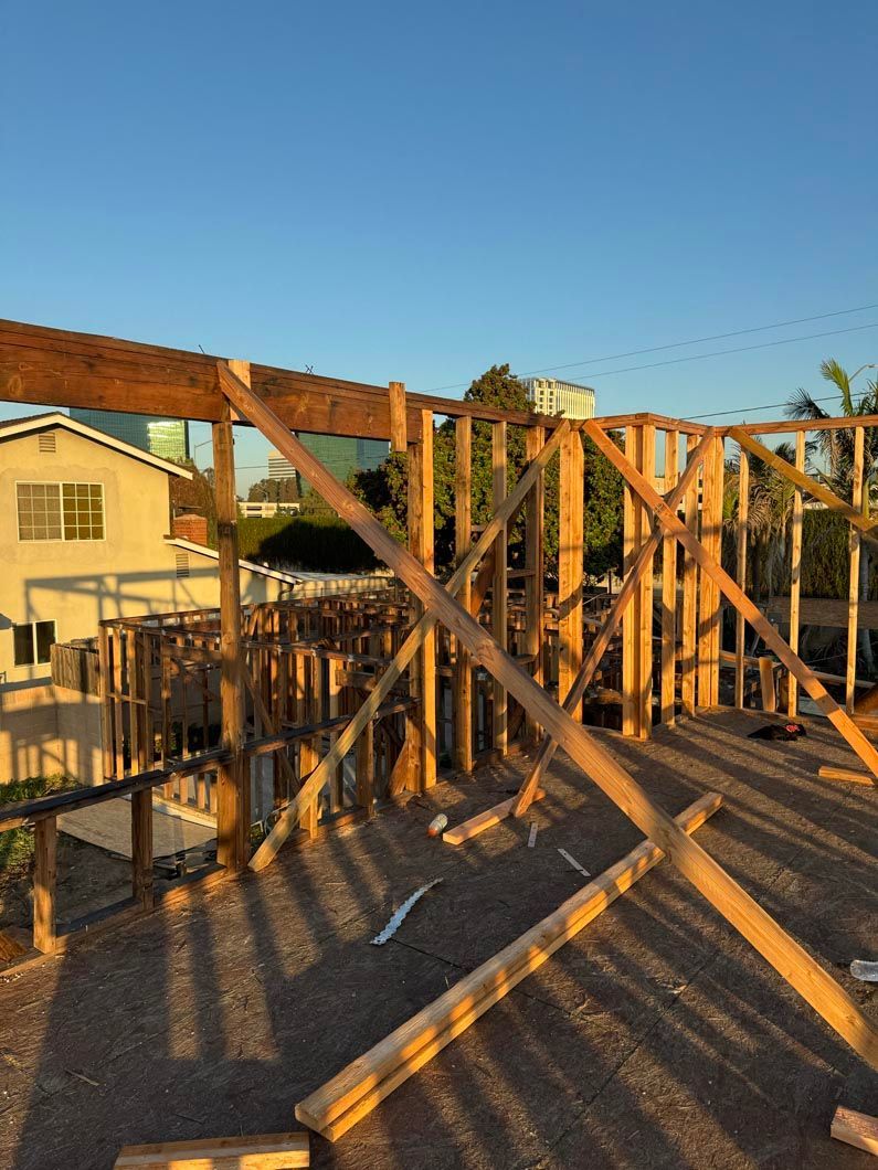 Wooden house frame under construction at sunset, with beams, supports, and neighboring homes visible.