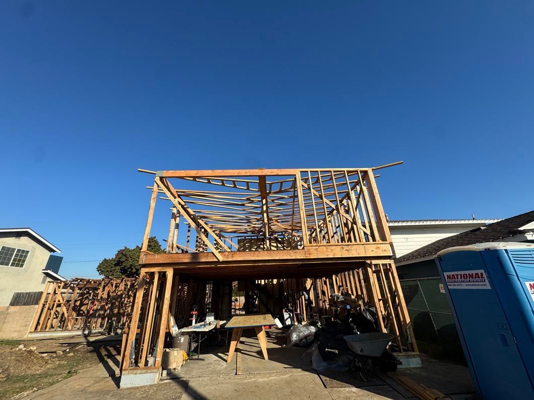 Partially built wooden deck framing beside a house, with scaffolding and construction materials under a blue sky