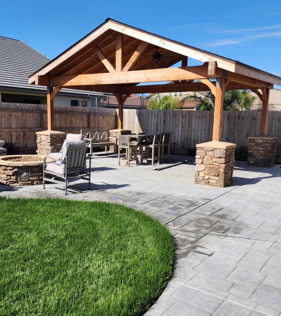 Backyard patio with wooden pergola, stone columns, outdoor dining set, and curved grass lawn.