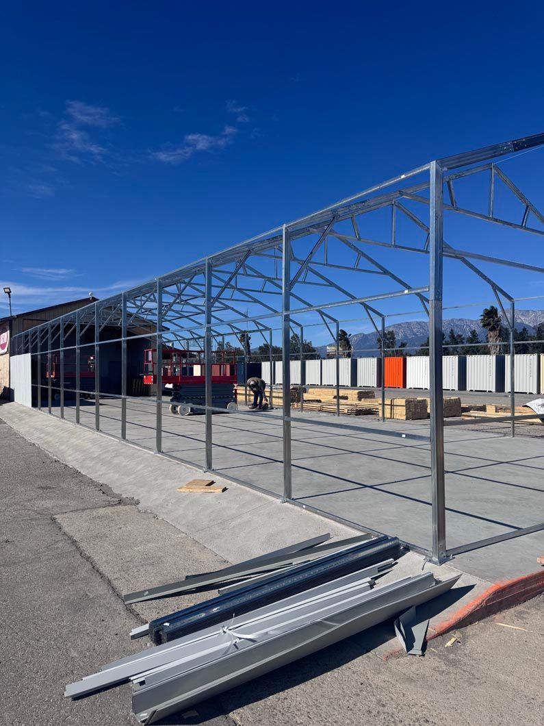 Steel-frame structure under construction beside a paved area, with metal beams stacked in the foreground.