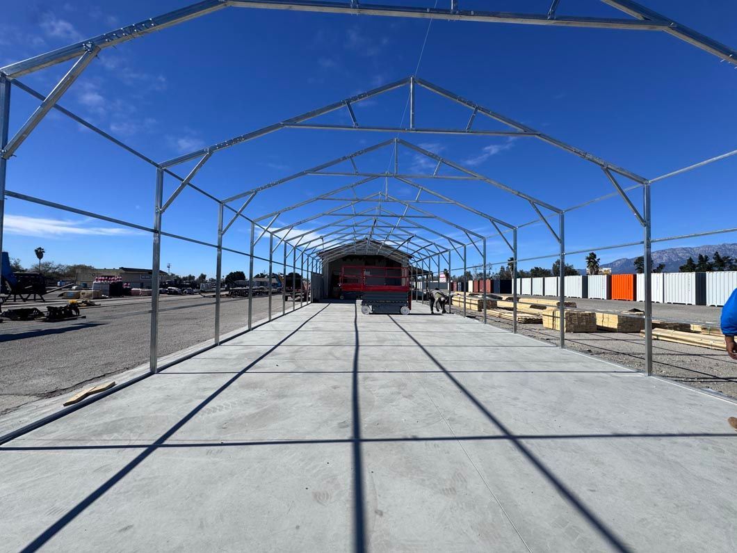 Concrete path under a metal frame structure on a sunny day, with open ground and buildings in the distance