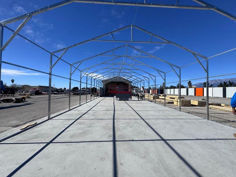 Empty concrete lot with a metal frame canopy under construction and a red building in the distance.