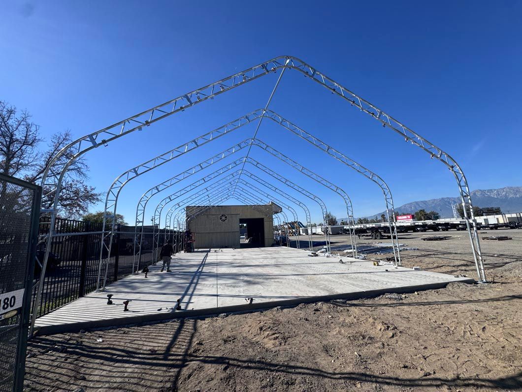 Outdoor event tent frame over a concrete pad under a clear blue sky, with a small building in the background