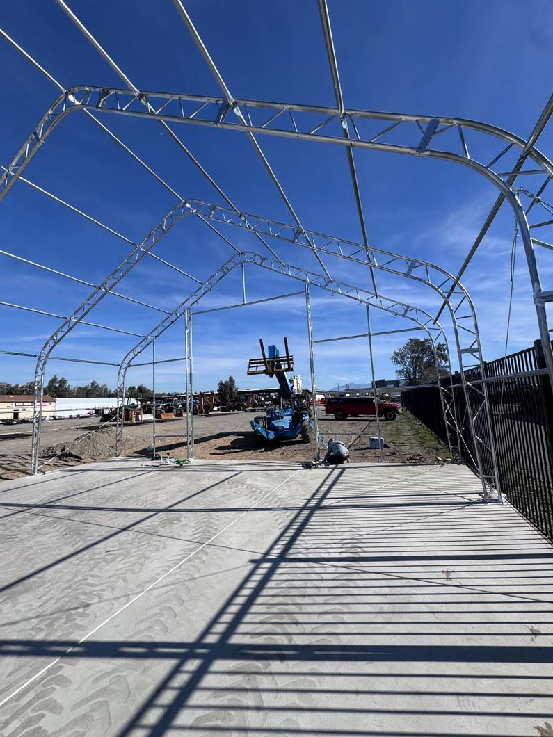 Steel frame structure under construction on a sunny industrial site, with a blue lift in the center.