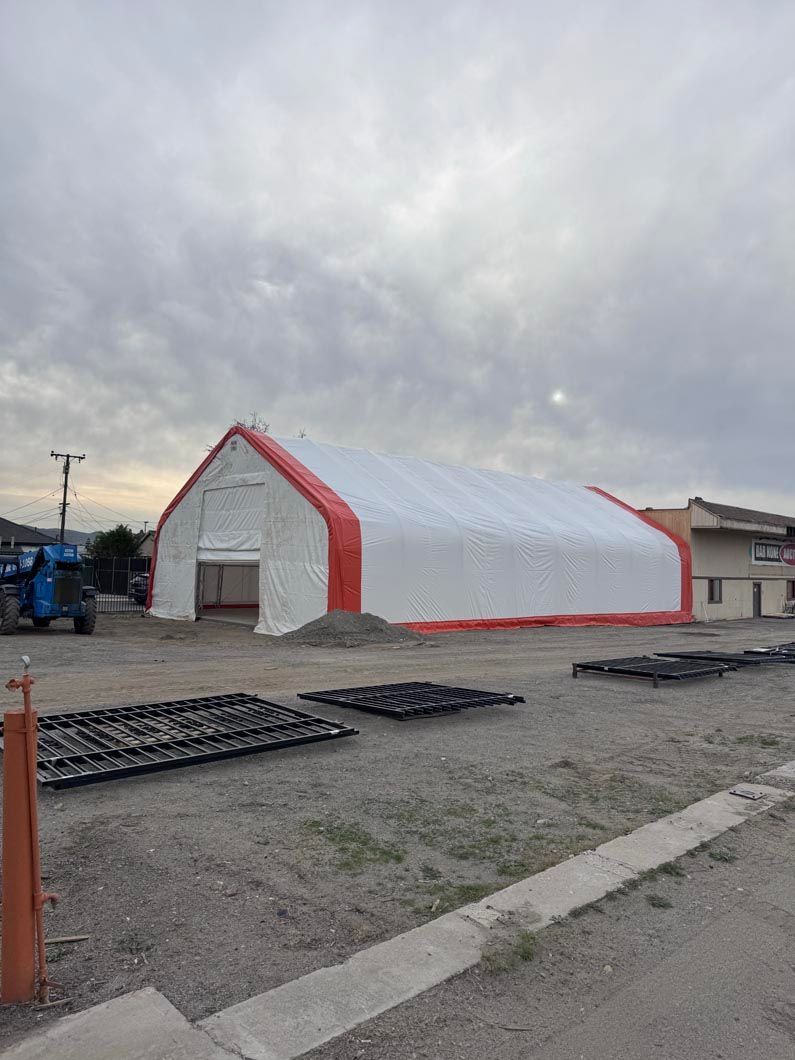 White arched tent building with red trim on gravel lot under cloudy sky, with pallets in front