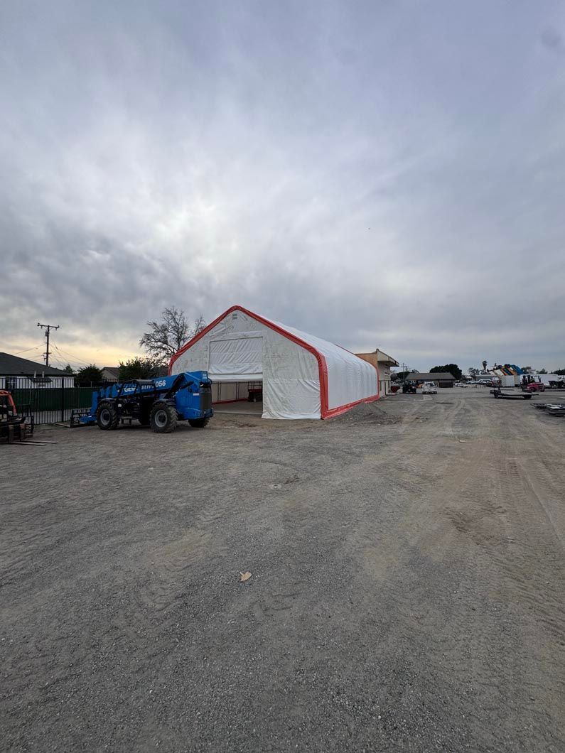 Blue tractor beside a white tent with red trim on a gravel lot under a cloudy sky