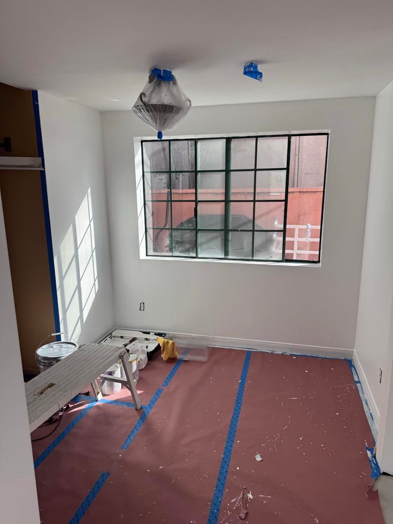 Small room under renovation with taped floor, white walls, window, and hanging light fixture.