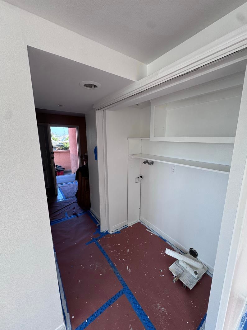 Small unfinished closet or hallway with white shelving, a white door, and red floor protected by blue tape.