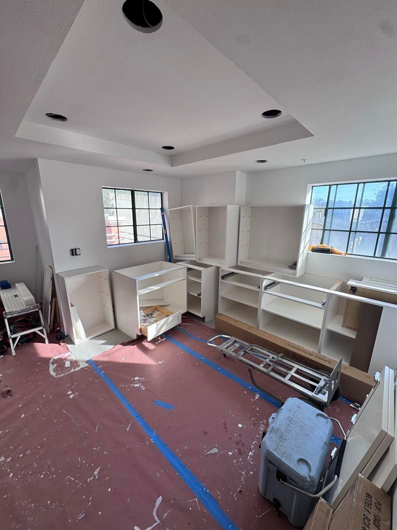 Unfinished kitchen with white cabinets, red floor covering, and windows letting in daylight.