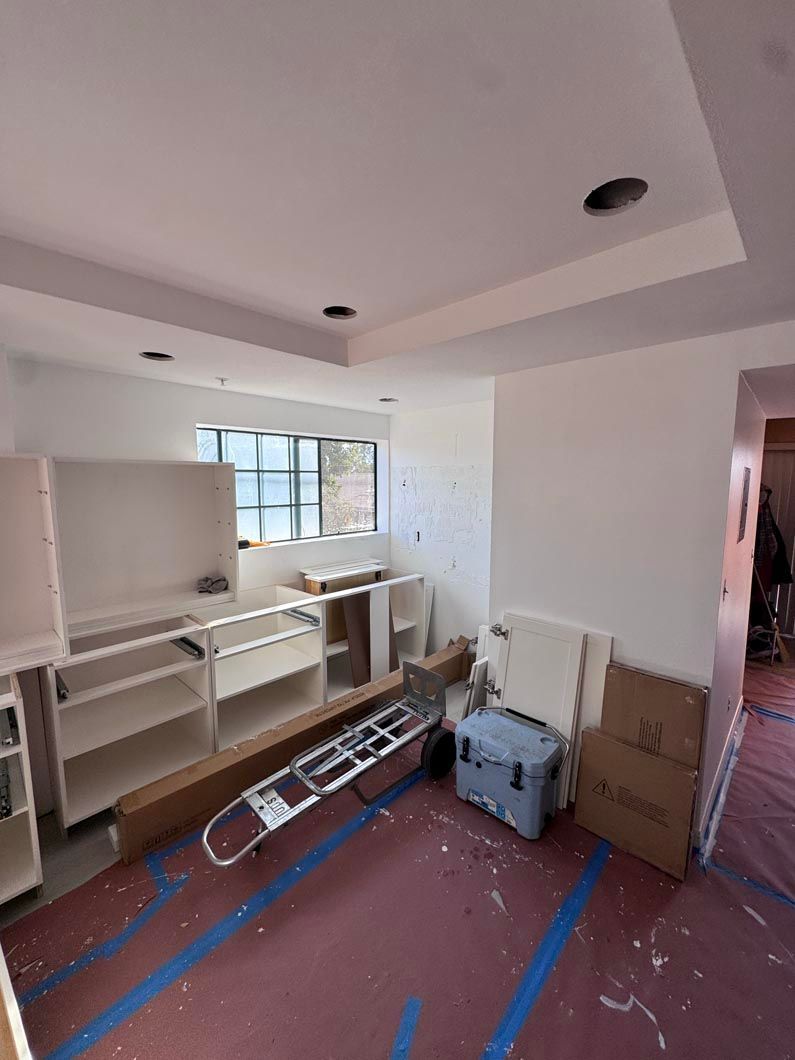 Unfinished room with white cabinets, window, ladder, boxes, and blue tape on the floor and ceiling.